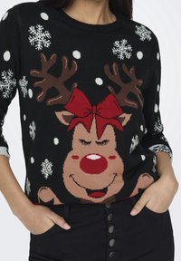 Svart stickad tröja med en leende ren med röd näsa, röd rosett och bruna horn, accentuerad med vita snöflingor och prickar.