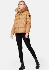 Beżowa kurtka puchowa z wysokim kołnierzem, z poziomą pikowaną fakturą i okrągłą czarną łatą na rękawie. Noszona z czarnymi legginsami.