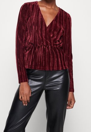 Frau trägt eine burgunderrote, langärmelige Wickelbluse aus Samt und schwarze, hoch taillierte Lederhosen, steht mit entspannten Armen.