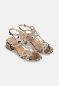 Sandalias de tacón beige con un intrincado diseño troquelado y acentos de strass. Presentan un acabado metálico y una correa al tobillo con hebilla.