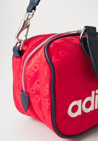 Rød nylonveske med subtilt adidas-logo mønster, hvitt brodert logo, sølv glidelås, og en marineblå stropp og detaljer. Rektangulær form.