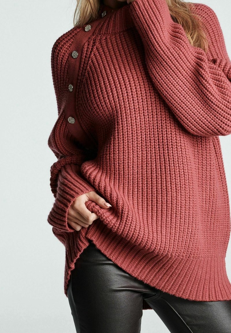 Femme portant un pull surdimensionné côtelé de couleur vieux rose avec des boutons métalliques à l'épaule et un pantalon en cuir noir, la main agrippant l'ourlet du pull.