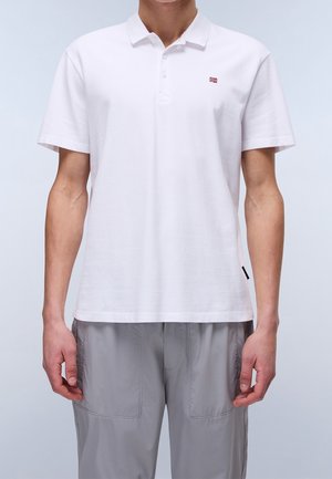 Mann trägt ein unifarbenes weißes kurzärmeliges Poloshirt mit einem kleinen Flaggenlogo auf der Brust und hellgraue Hosen.