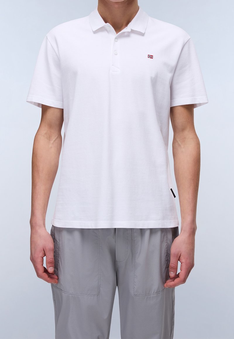 Homme portant un polo blanc uni à manches courtes avec un petit logo de drapeau sur la poitrine et un pantalon gris clair.