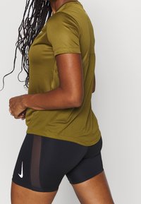 Olivegrön atletisk tröja med korta ärmar, med en ventilerande meshdesign, ihop med svarta åtsittande shorts prydda med en vit logotyp.