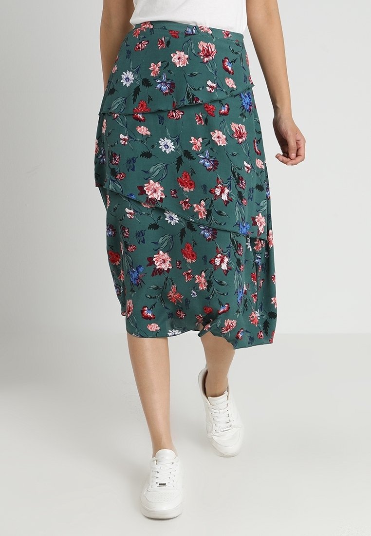 Mönstrad midikjol i mörkgrön tyg med lagerdesign, prydd med röda, rosa och blå blommor, som matchas med vita sneakers.