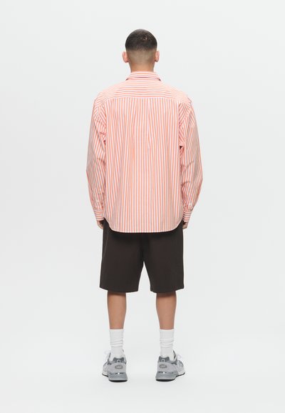 Man standing facing away wearing a pink and white striped long-sleeve shirt, black shorts, white socks, and gray sneakers.