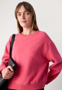 Pink sweatshirt with a round neckline, ribbed cuffs, and hem. Features an embroidered logo on the chest. Model holds a black handbag.