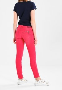 Persona vestida con pantalones ajustados rosados brillantes, una camiseta negra de manga corta y zapatillas blancas con estampados de animales, de pie mirando hacia atrás.