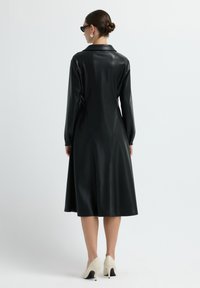 Žena v černých midi šatech s dlouhým rukávem z umělé kůže s límečkem, oblečená v bílých vysokých podpatcích, slunečních brýlích, se stříbrnými kruhovými náušnicemi a vlasy svázanými do bun.