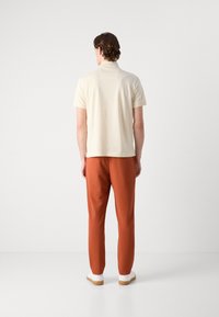 Kremfarget kortermet polo skjorte kombinert med rustfargede teksturerte bukser og hvite sneakers, sett bakfra.