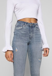 Camisa blanca de manga larga y corta con puños acampanados, combinada con jeans de tiro alto en azul claro que presentan áreas desgastadas en los muslos.