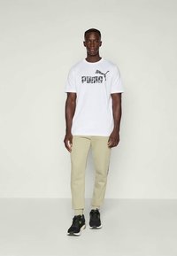 Camiseta blanca con un logo "PUMA" en camuflaje negro, combinada con pantalones beige claros y zapatillas deportivas negras. Diseño simple y ajuste casual.