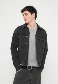 Črna denim jakna z metalnimi gumbi, opremljena z dvema prsnikoma in klasičnim ovratnikom, nošena čez sivo majico z okroglim ovratnikom.