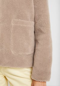 Beige fleecejakke med en blød tekstur, der har en lomme og en afslappet pasform, vist med lysegule bukser.