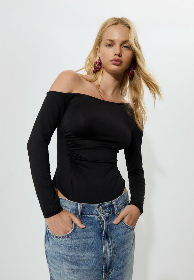 Svart off-the-shoulder bodysuit med långa ärmar och samlade sidor, ihopparat med ljusblå högmidjade jeans.