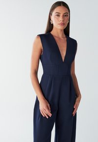 Temno modra brezrokavna kombinezon s globokim V-izrezom, strukturiranim zgornjim delom in širokimi hlačnicami. Gladek material z nežnimi teksturnimi detajli.