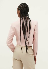 Jasnoróżowa krótka marynarka z długimi rękawami, o strukturalnym kroju. Noszona z beżowymi spodniami, prezentująca prosty i czysty styl.