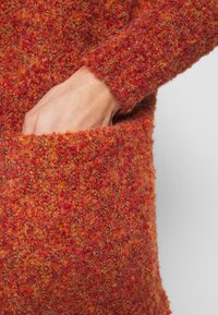 Sweter w kolorze rdzy z dzianiny o fakturowanym wzorze. Posiada kieszeń z przodu oraz ściągacz na rękawach. Miękki, puszysty materiał.