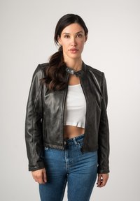 Schwarze Lederjacke mit rundem Kragen, ausgestattet mit metallischen Druckknöpfen und einem vorderen Reißverschluss. Getragen über einem weißen, gekürzten Tanktop und blauen Jeans.