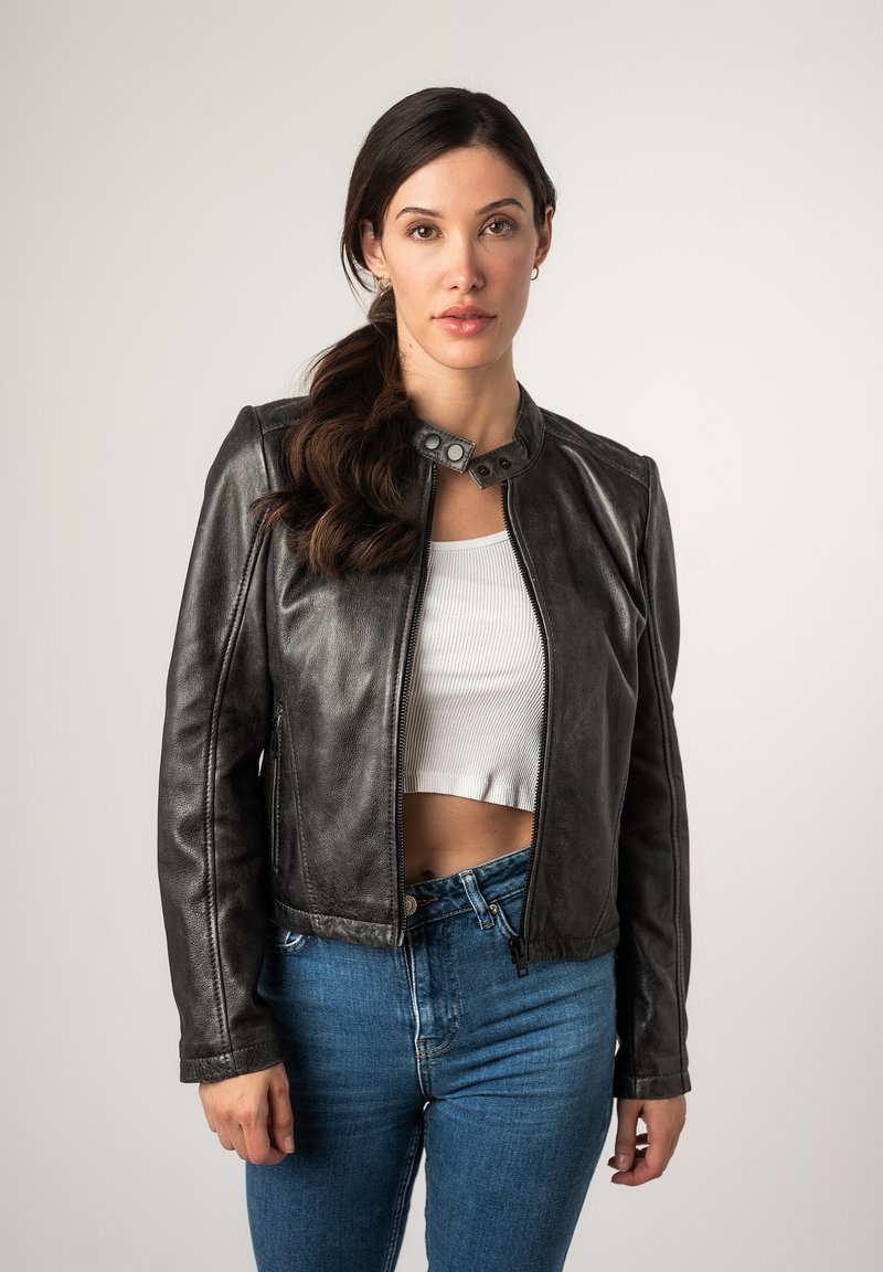 Schwarze Lederjacke mit rundem Kragen, ausgestattet mit metallischen Druckknöpfen und einem vorderen Reißverschluss. Getragen über einem weißen, gekürzten Tanktop und blauen Jeans.