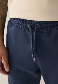 Tengerészkék színű melegítőnadrág texturált derékpánttal, ezüst színű fém részletekkel ellátott zsinórral. Sima, rugalmas anyagból készült.