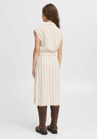Cremfarbentes Kleid mit vertikalen marineblauen Streifen, kurzen Ärmeln, Taillengürtel und Schlitzsaum, kombiniert mit braunen kniehohen Stiefeln. Glatte Textur.