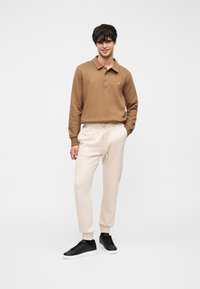 Καφέ μακρυμάνικη πόλο μπλούζα με γιακά και χρυσό λογότυπο, σε συνδυασμό με μπεζ παντελόνι jogger και μαύρα αθλητικά παπούτσια. Ομαλή υφή συνολικά.