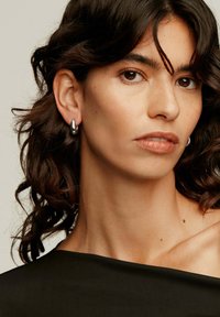 Pendientes de aro plateados llevados en la oreja izquierda de una persona con cabello oscuro, ondulado y de longitud hasta los hombros, que presentan un acabado suave y reflectante.