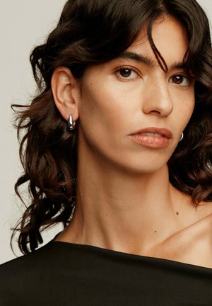 Pendientes de aro plateados llevados en la oreja izquierda de una persona con cabello oscuro, ondulado y de longitud hasta los hombros, que presentan un acabado suave y reflectante.