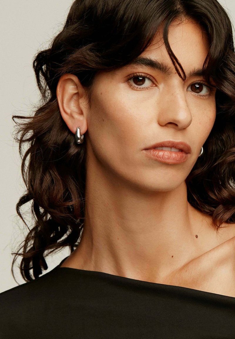 Pendientes de aro plateados llevados en la oreja izquierda de una persona con cabello oscuro, ondulado y de longitud hasta los hombros, que presentan un acabado suave y reflectante.