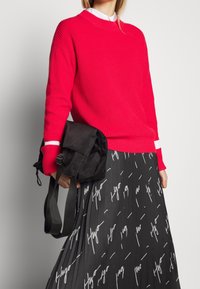 Persona con un suéter rojo brillante y una falda plisada negra con escritura blanca, sosteniendo un bolso negro plegado con correas frente a un fondo simple.