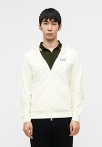 Κρεμ φούτερ με φερμουάρ, απαλής υφής, που διαθέτει κουκούλα, μπροστινές τσέπες και λογότυπο στο στήθος· συνδυασμένο με σκούρο πράσινο πόλο από κάτω.