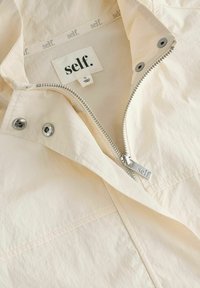 Chaqueta beige con cremallera metálica y tira grabada con "self.", botones de presión y etiqueta de la marca "self." en el interior del cuello, talla pequeña, fabricada en China.