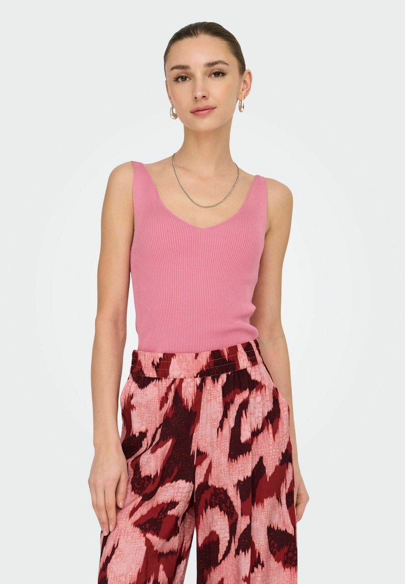 Femme portant un haut sans manches côtelé rose et un pantalon large taille haute rose et rouge foncé à motifs, debout devant un fond uni.