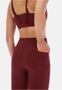 Sostén deportivo burdeos con tirantes ajustables y leggins a juego, que incluyen un pequeño bolsillo de acento naranja y detalles de tela texturizada.