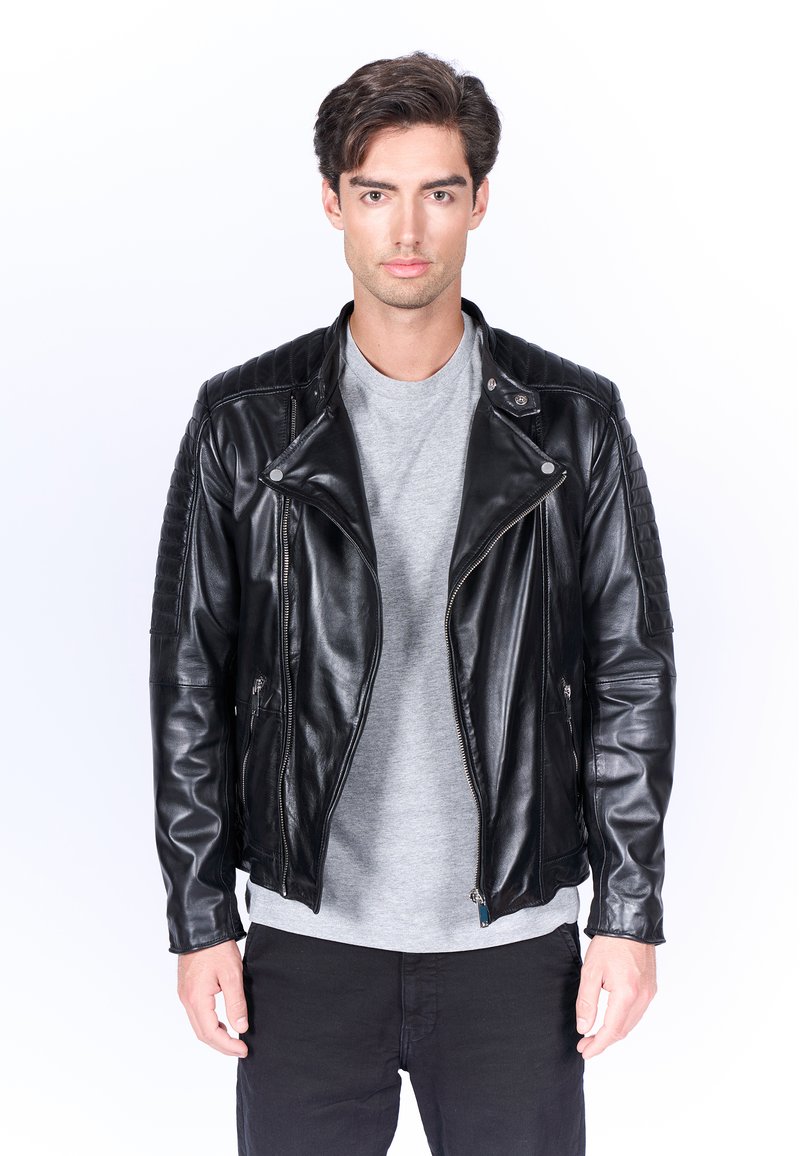 Giacca da moto in pelle nera con dettagli trapuntati sulle spalle, tasche con zip e hardware argento, abbinata a una maglietta grigia.