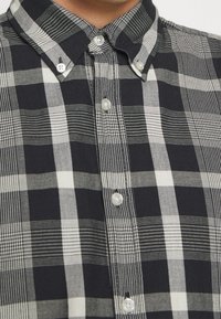 Koszula w kratę w czerni i bieli, z usztywnianym kołnierzem, perłowymi guzikami oraz różnymi wzorami kraty na teksturowanym materiale.