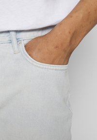 Jasnoniebieskie dżinsy z widoczną boczną kieszenią, ozdobione subtelnymi szwami i lekko zniszczonym wyglądem.