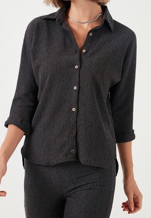 Femme portant une chemise boutonnée texturée foncée avec des manches retroussées et un pantalon assorti, montrée du cou aux hanches sur fond blanc.