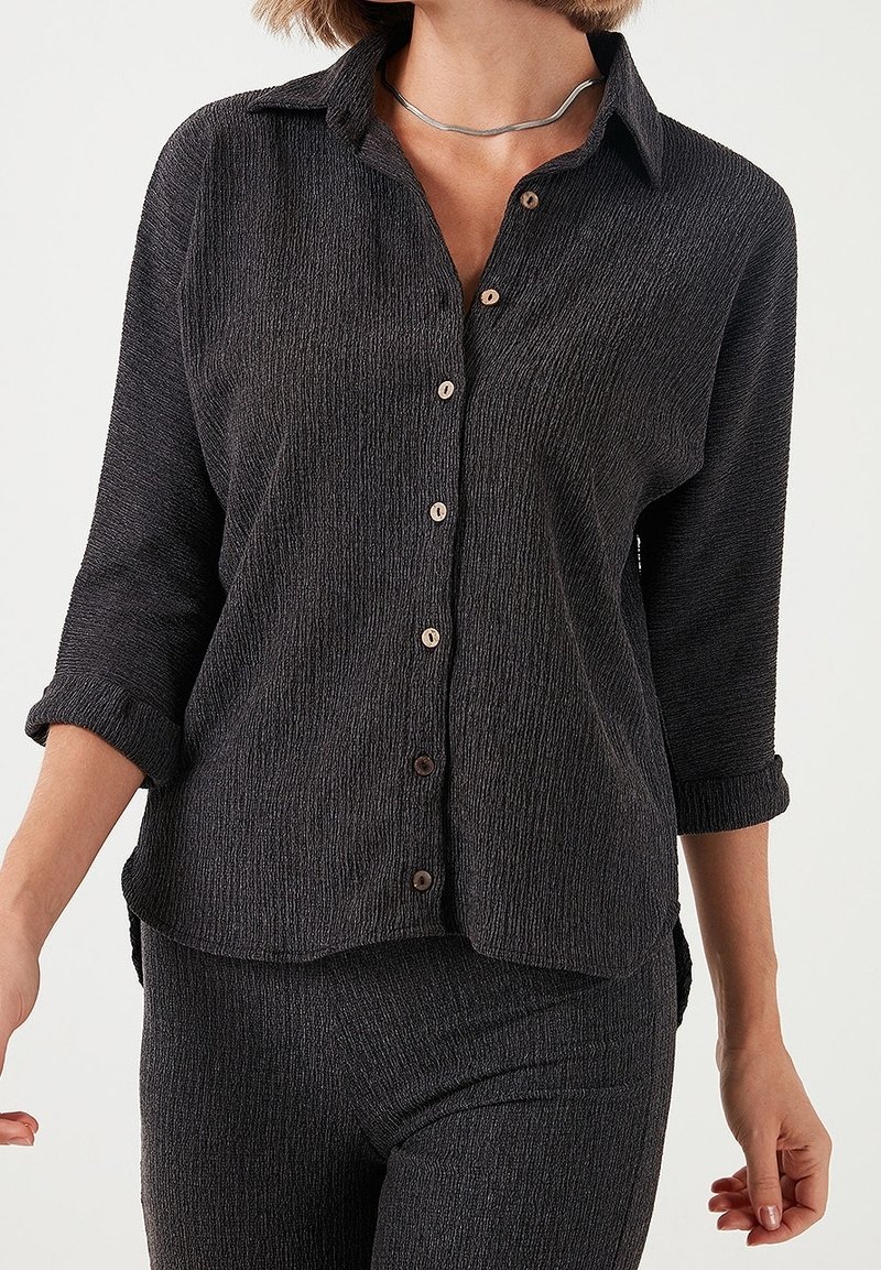 Femme portant une chemise boutonnée texturée foncée avec des manches retroussées et un pantalon assorti, montrée du cou aux hanches sur fond blanc.