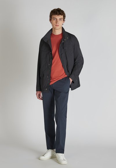 Giacca nera leggera con colletto alto, due tasche frontali, indossata sopra una maglietta color ruggine. Abbinata a pantaloni navy e sneakers bianche.