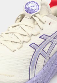 Witte sportieve sneaker met gestructureerde stof, paarse accenten, zichtbare veters en een ronde tag met het label "Tokyo Collection."