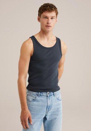 Jonge man met kort, krullend haar, gekleed in een marineblauw mouwloos T-shirt en lichtblauwe jeans op een effen beige achtergrond.