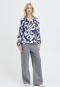 Langärmelige Bluse mit abstraktem blau-cremefarbenem Muster, entspannter Passform und elastischen Bündchen, kombiniert mit grauen Jeans mit weitem Bein.