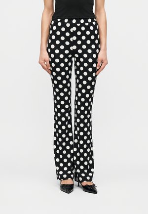 Femme portant un pantalon noir à jambes larges avec de grands pois blancs, des talons noirs à bout pointu et un haut sans manches noir, debout droite.