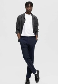 Σκούρο γκρι πουκάμισο με κουμπιά πάνω από λευκό μπλουζάκι, συνδυασμένο με ναυτικά παντελόνια και μαύρες λοφάδες. Λείες υφές, απλός σχεδιασμός και ελάχιστες λεπτομέρειες.