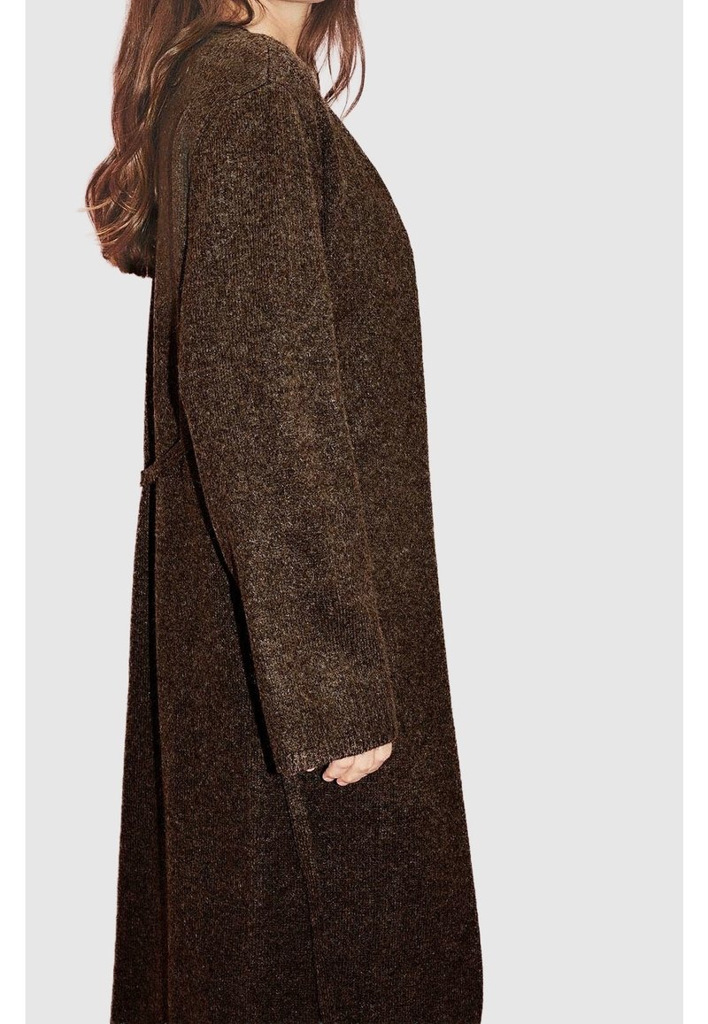 Femme portant un long cardigan en maille brun foncé texturé à manches longues, debout de profil devant un fond clair uni.