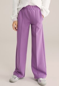 Cilvēks, kurš valkā augsta jostasvietas platu staru lavandas krāsas bikses ar priekšējām šuvēm, baltu krekliņu ar garām piedurknēm un baltus tenisa apavus. Rokas ieliktas kabatās.