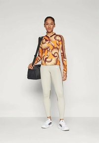 Dlhý rukáv športový top s oranžovým a žltým perím, skombinovaný so svetlo sivými legínami a bielymi teniskami. Model drží čiernu tašku.